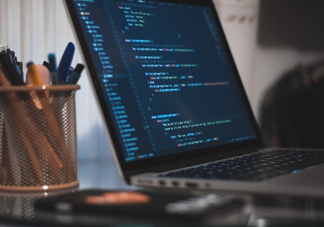 Image shows a laptop monitor on a desk with the screen filled with lines of code